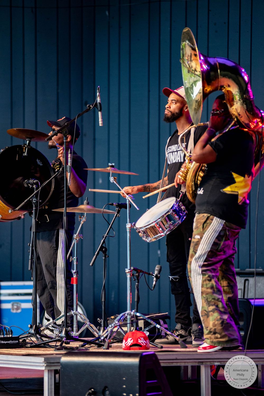 Live band performs on stage with drummer, guitarist, and tuba player under colorful lights at outdoor concert.