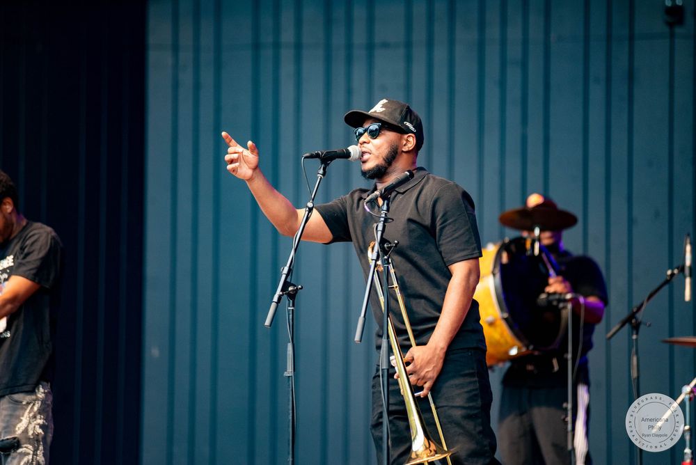A musician plays trombone and speaks into a microphone on an outdoor stage during a live music performance.
