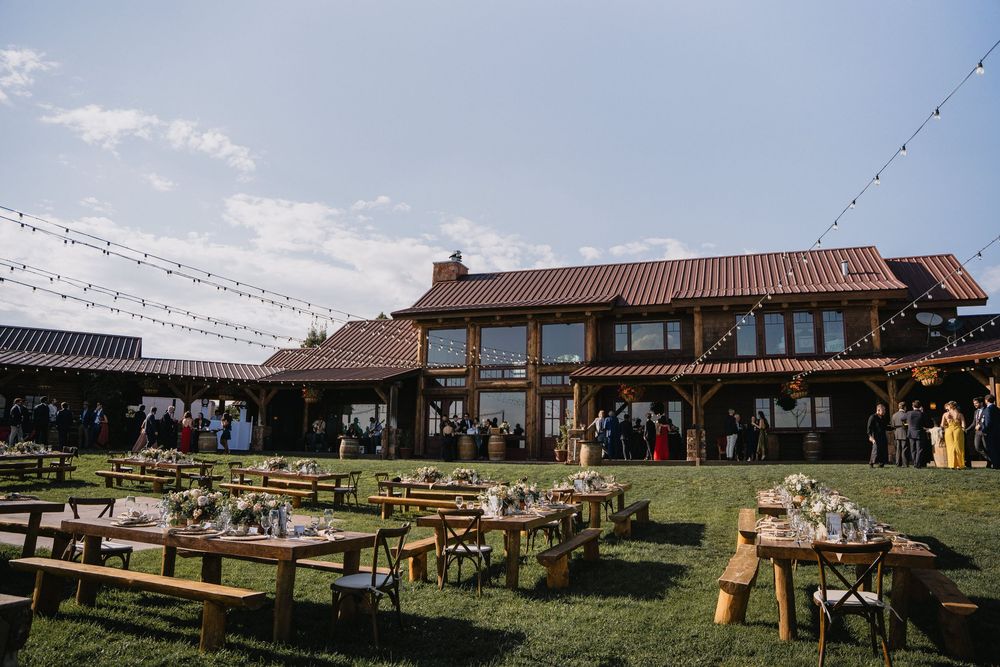 Rustic barn wedding venue with outdoor reception tables on green lawn, string lights, and guests mingling under blue sky.