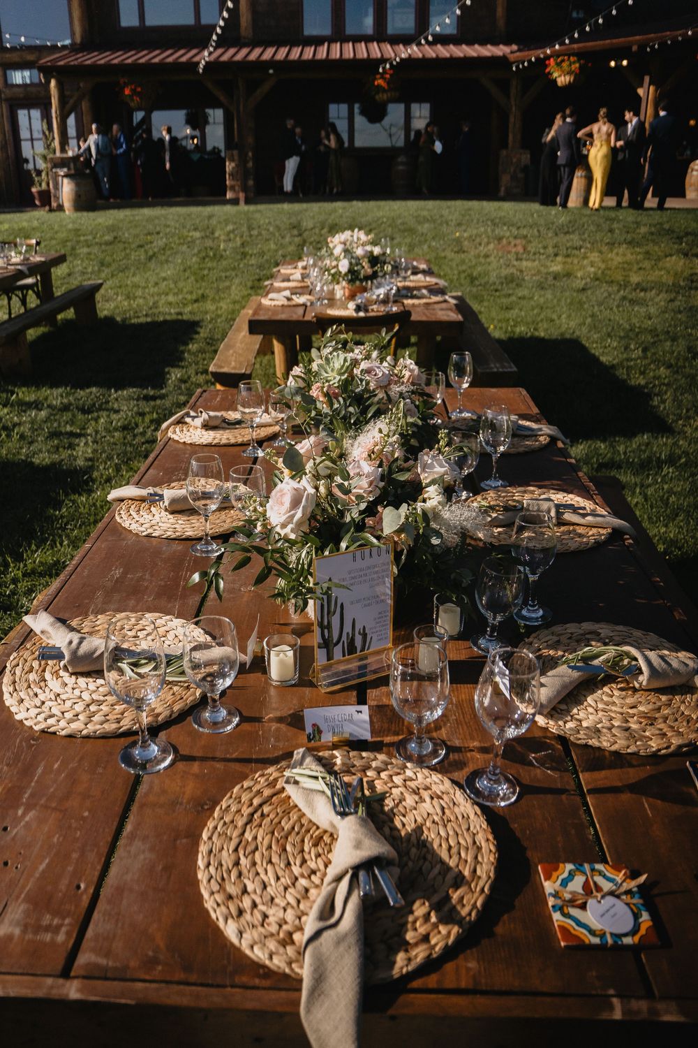 Rustic outdoor wedding reception with long wooden farm tables, woven chargers, floral centerpieces, and string lights near a barn.