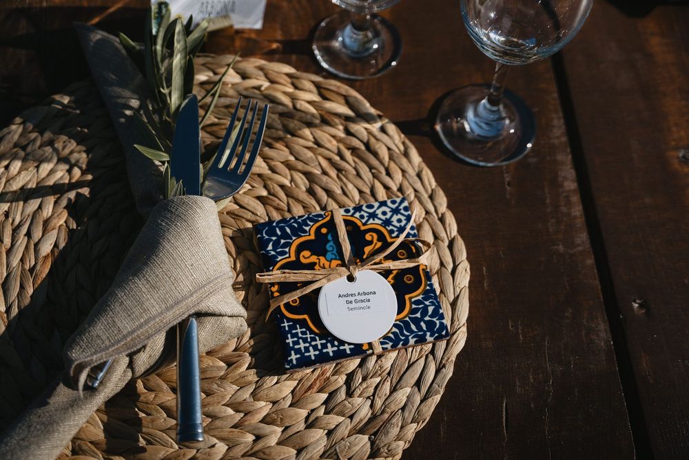 Elegant wedding place setting with woven charger, linen napkin, silverware, and blue floral favor on rustic wood table.