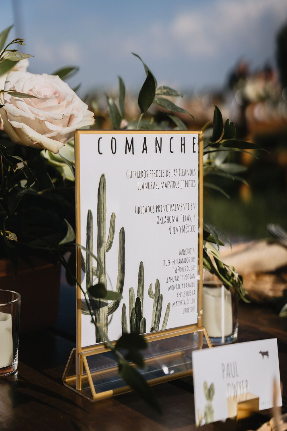 Gold-framed Comanche table sign with cactus illustration at a rustic wedding reception with floral arrangements.