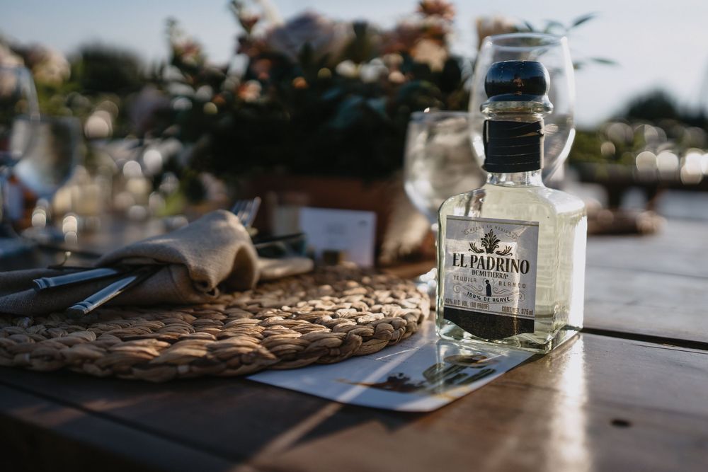 El Padrino tequila bottle on rustic wooden table with woven placemat and floral centerpiece at outdoor event.