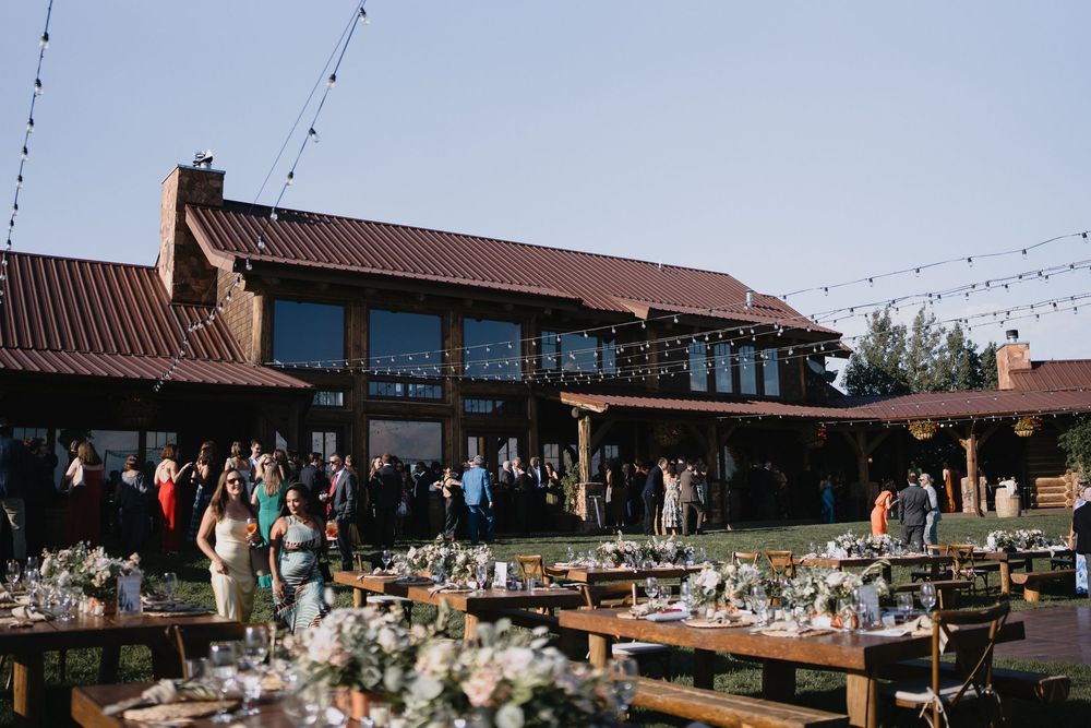 Outdoor wedding reception at rustic barn venue with string lights, wooden tables, floral arrangements, and guests gathered on green lawn.