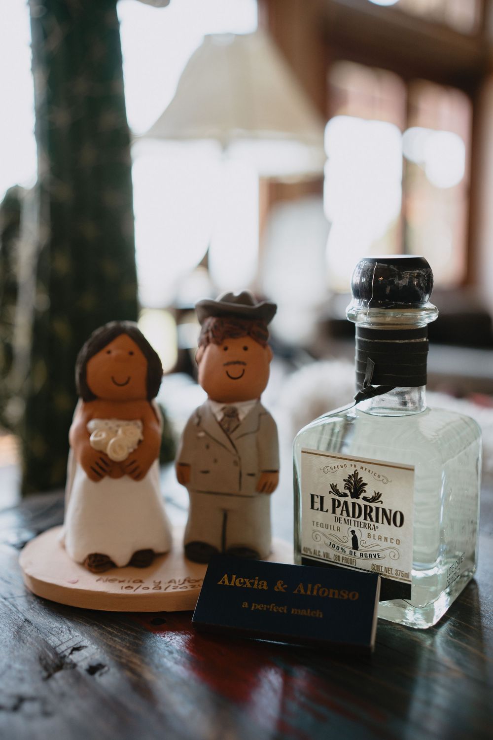 Wedding cake toppers beside El Padrino tequila bottle and Alexia & Alfonso name card on rustic wooden table.