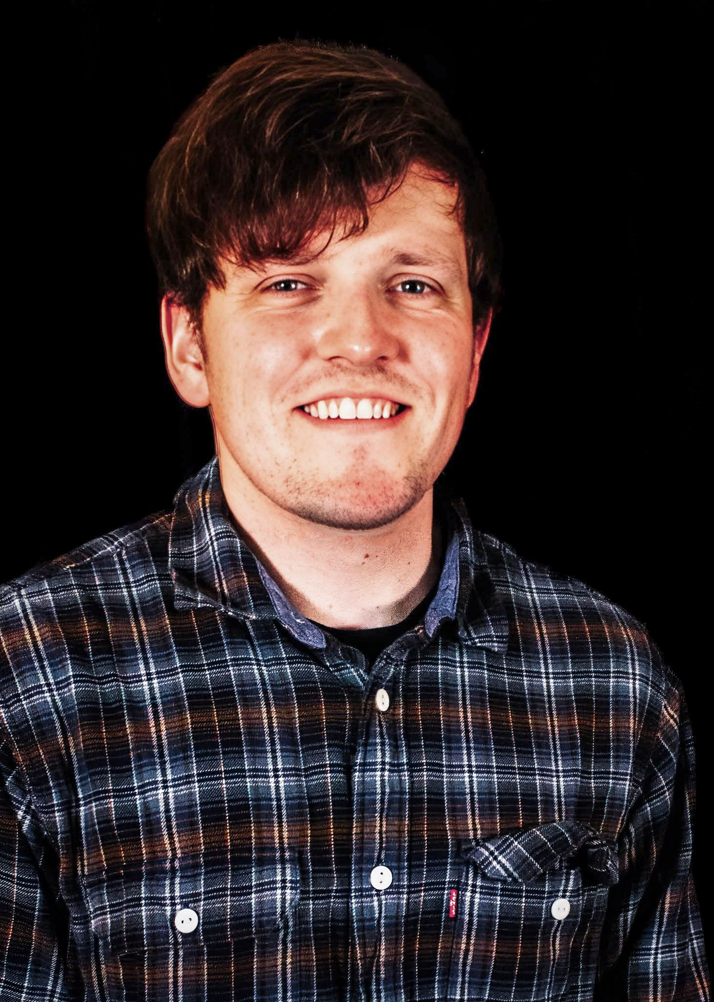 Smiling young man with brown hair wearing a plaid flannel shirt against a black background.