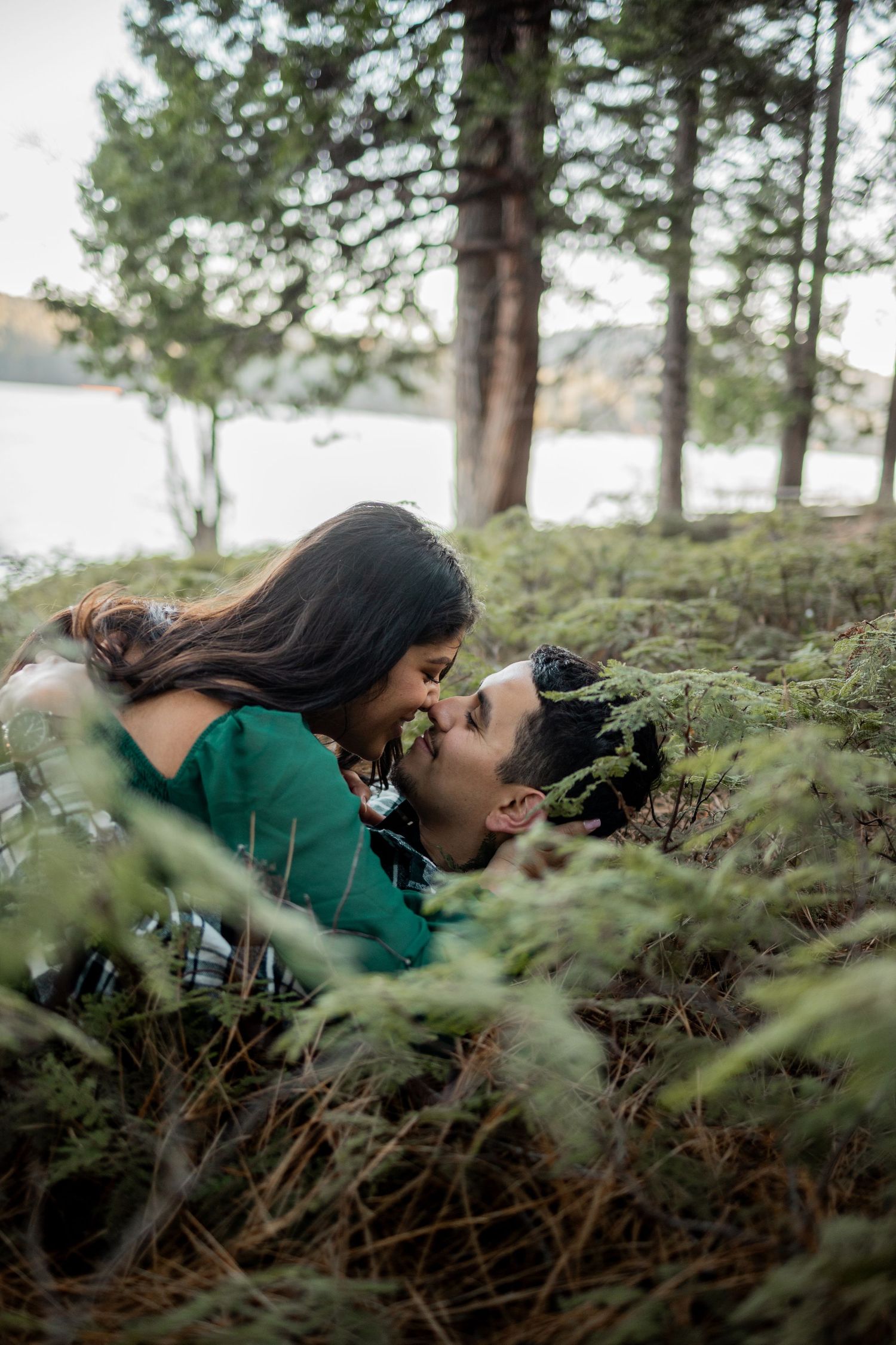 Couple sharing a romantic moment lying among lush green forest ferns near a serene lake.