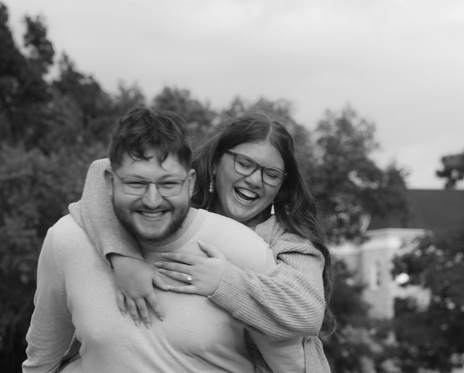 Black and white photo of a laughing couple, woman hugging man from behind outdoors with trees in background.