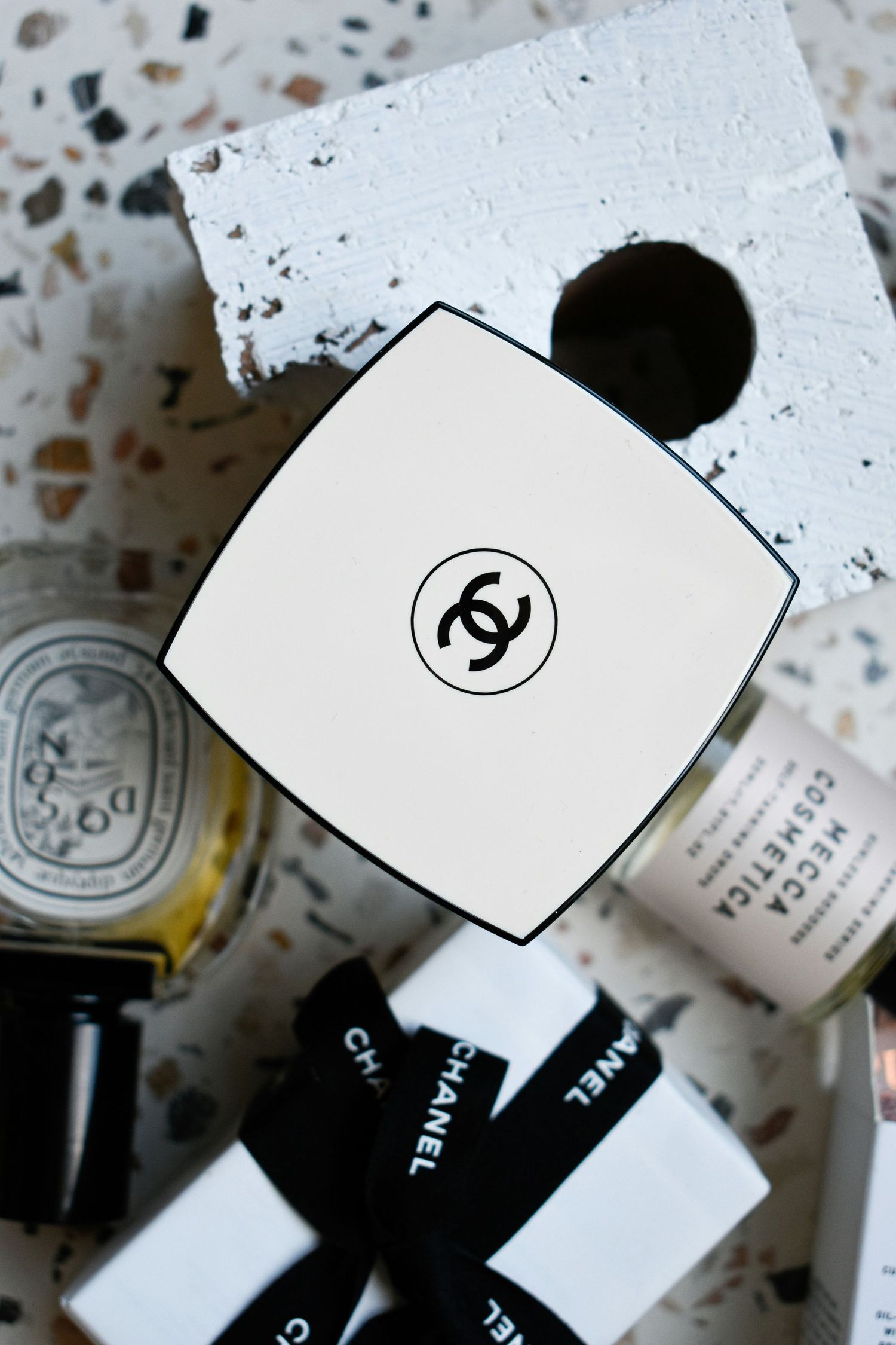 Chanel compact powder and branded boxes arranged on a terrazzo surface with Diptyque and BECCA cosmetics.