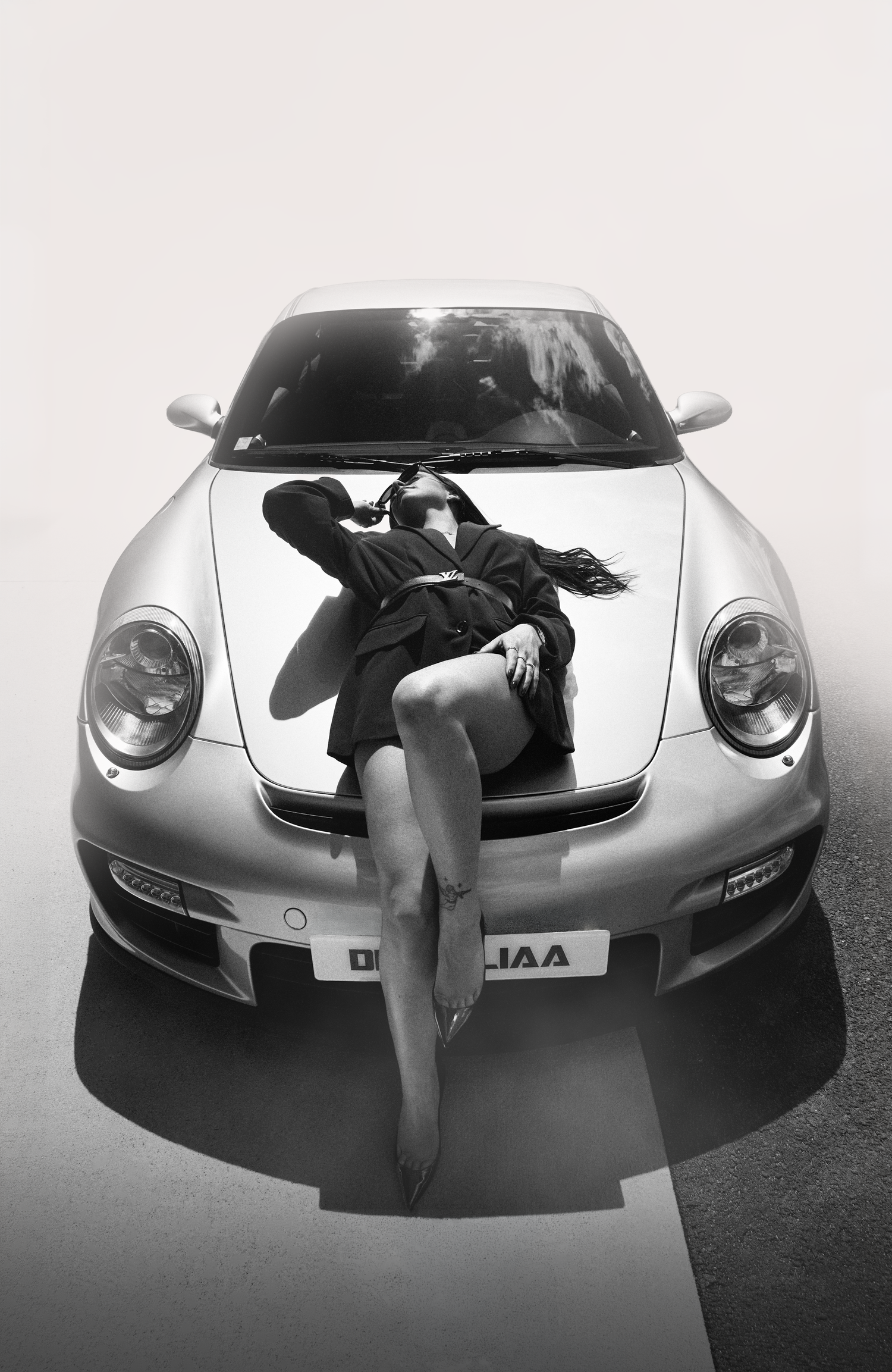 Black and white photo of a woman posing on the hood of a silver Porsche sports car, shot from above.