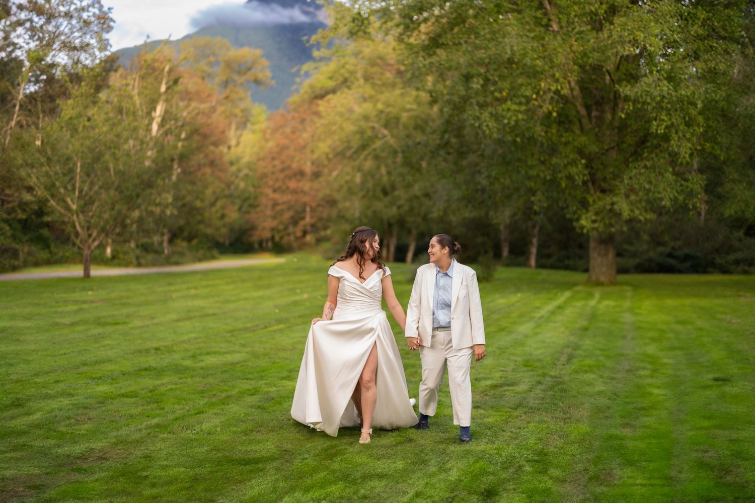Two smiling grooms in vests embrace outdoors among green trees on their wedding day.
