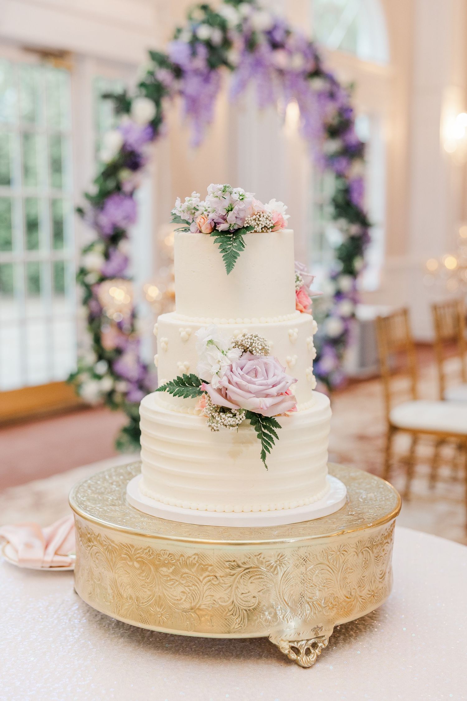 Three-tier white wedding cake with pink and lavender roses on gold stand, purple floral arch backdrop.