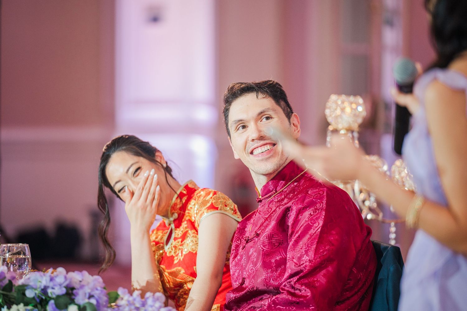 A smiling Asian couple in traditional red Chinese wedding attire laugh joyfully at their wedding reception.