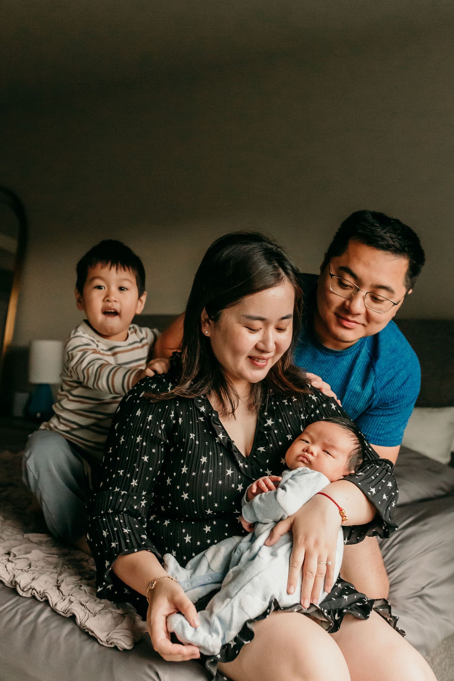 Asian family of four on bed, mother holding newborn as father and toddler smile warmly.