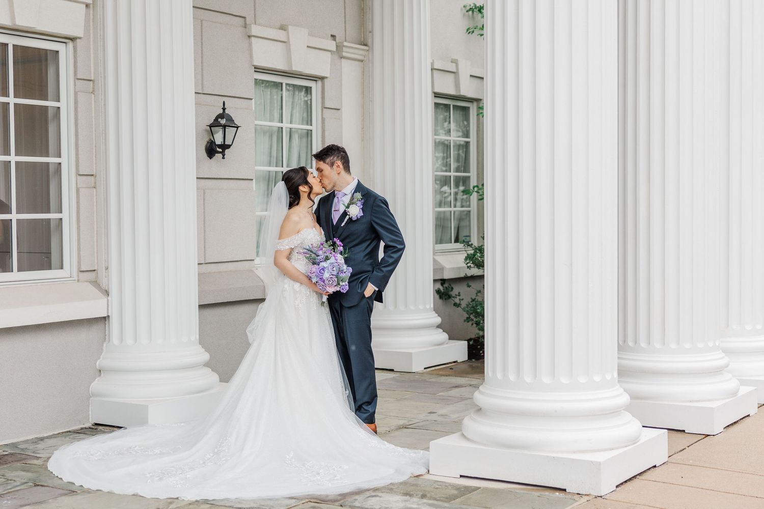 Bride and groom share a romantic kiss between white columns at an elegant wedding venue.