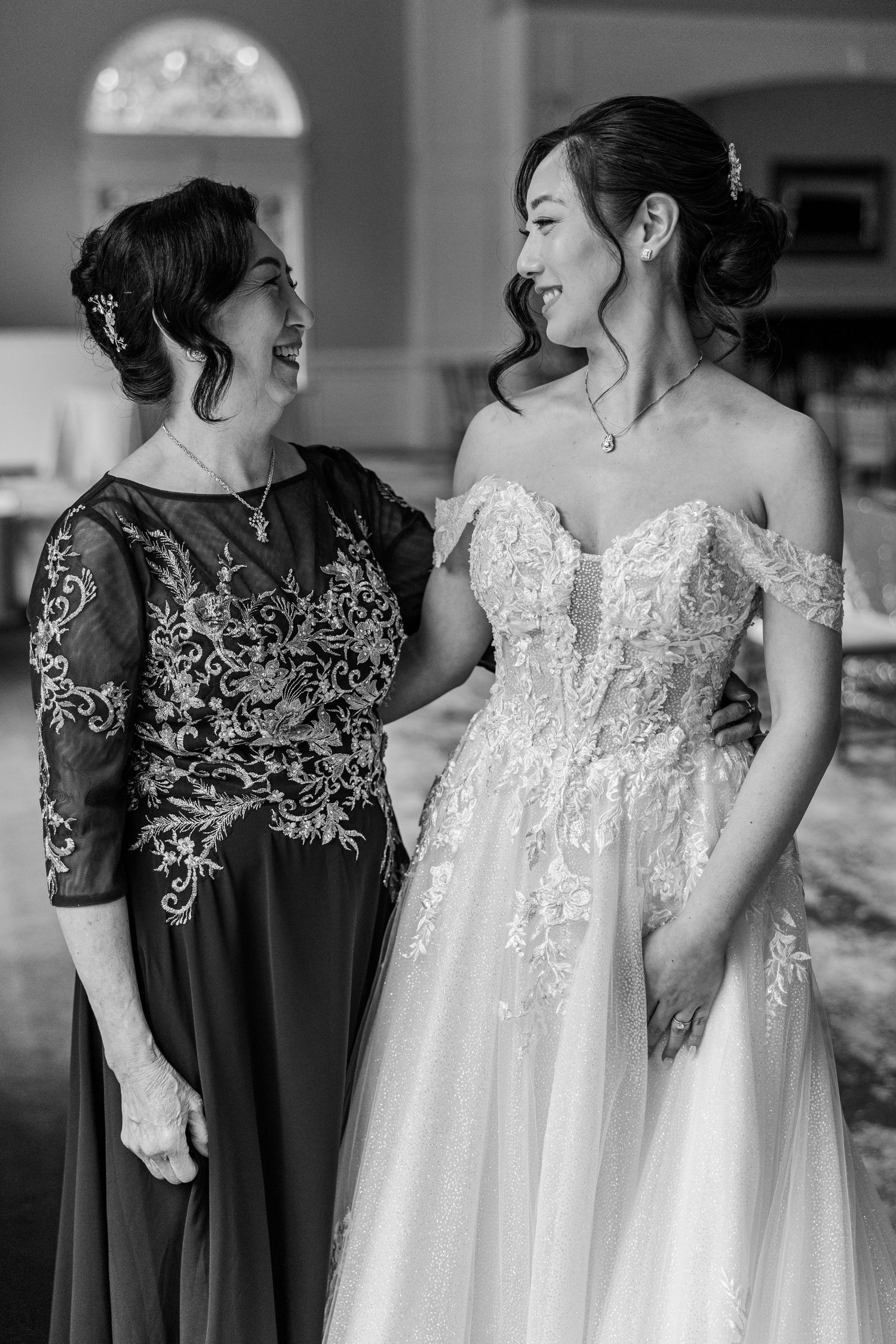 Black and white photo of a bride in lace off-shoulder gown smiling with an older woman in elegant dark dress.