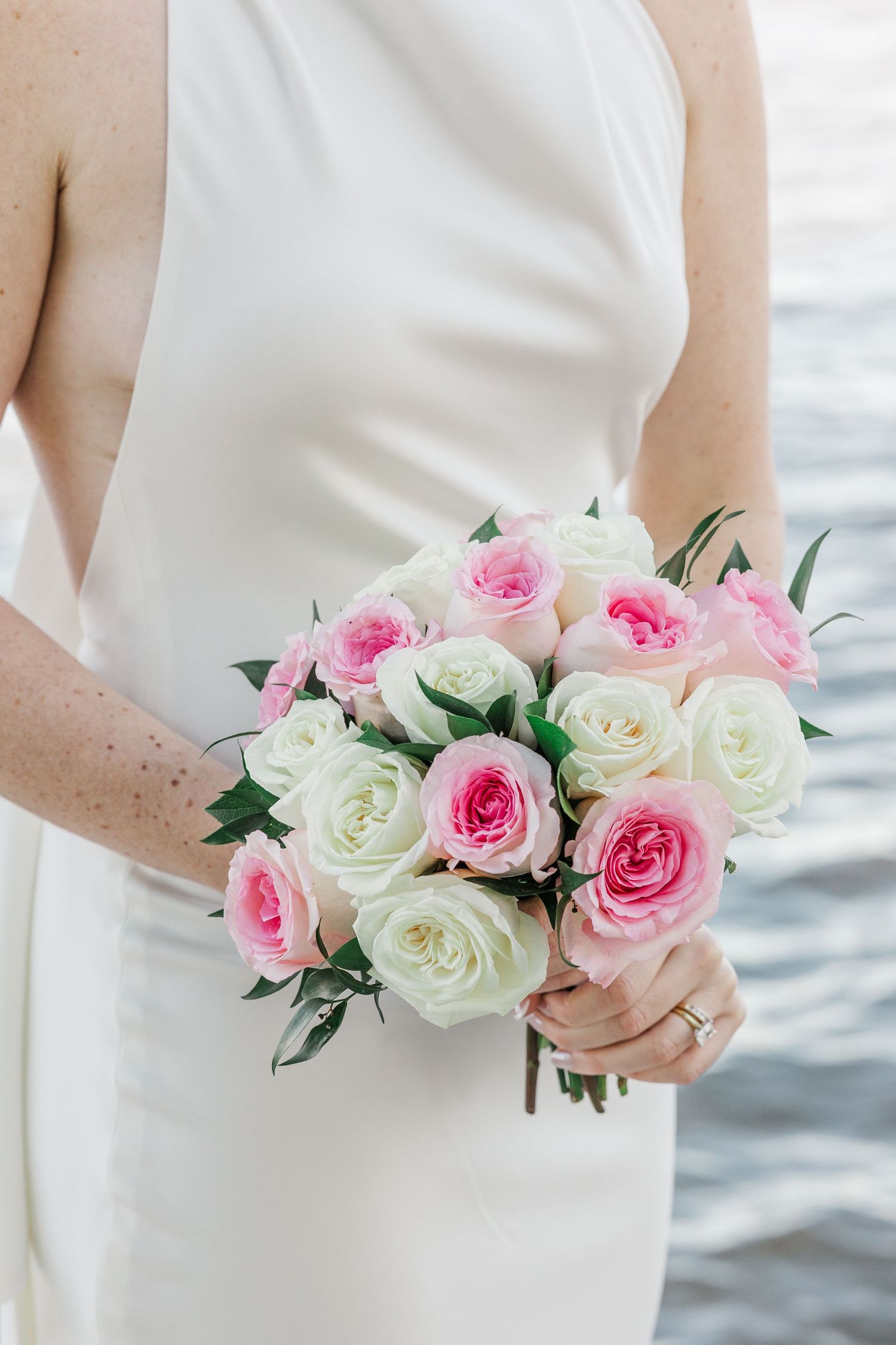 Bride in white dress holding a bridal bouquet of pink and white roses near water.