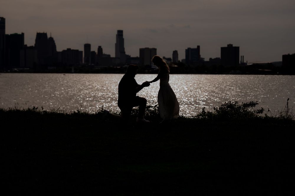 Silhouette of a couple at sunset by a city waterfront, man on one knee proposing to woman.