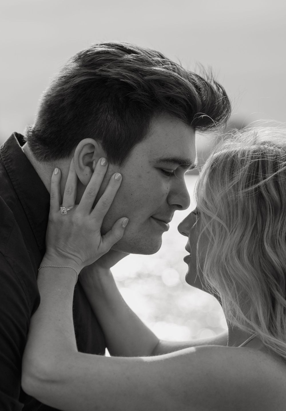 Black and white romantic couple almost kissing, woman wearing engagement ring tenderly holding mans face outdoors.