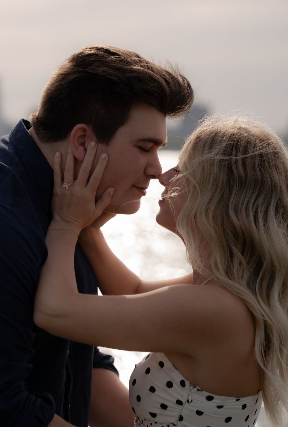Couple sharing a romantic almost-kiss moment outdoors near water, woman holding mans face tenderly.