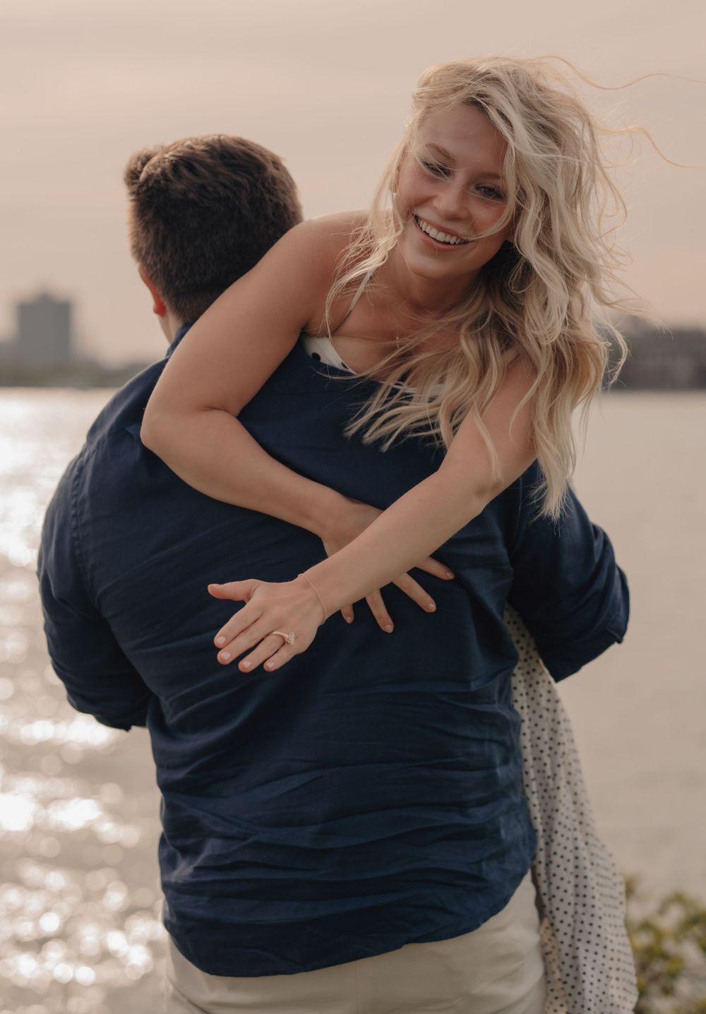 Smiling blonde woman being lifted by man in navy shirt near waterfront during romantic outdoor photo session.