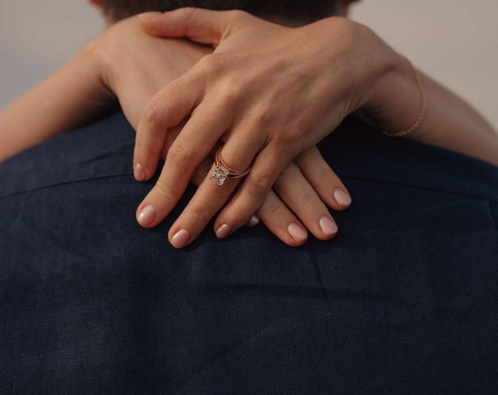 Close-up of a woman's hand with an engagement ring resting on a man's back in a warm embrace.