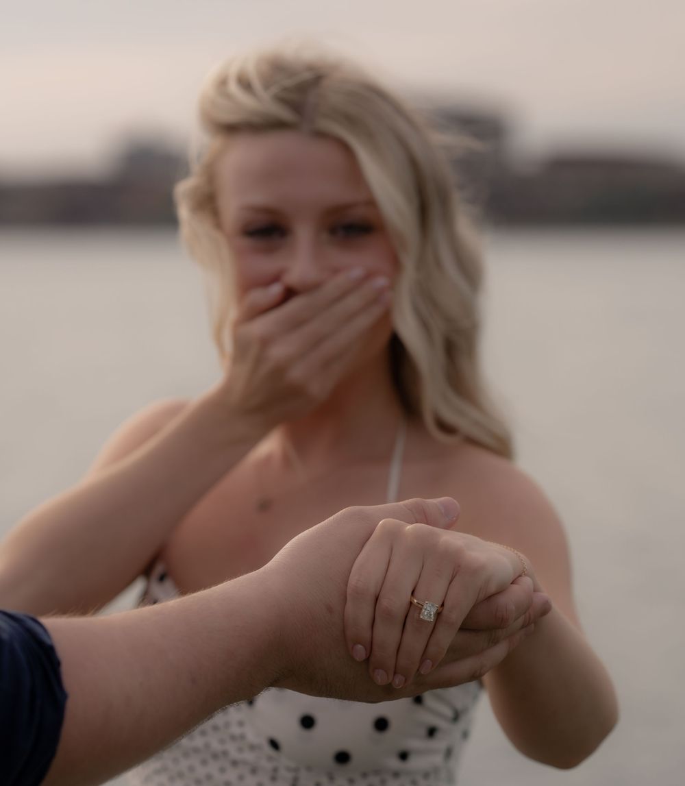 Emotional woman covers mouth in surprise as man slides diamond engagement ring onto her finger by waterfront.