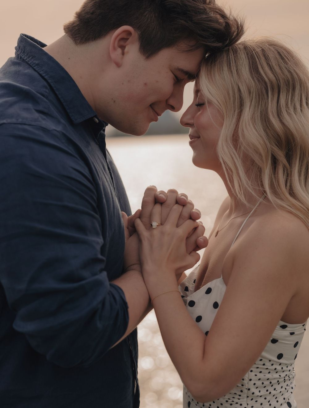 Romantic couple holding hands and touching foreheads tenderly outdoors, woman in polka dot dress, man in navy shirt.