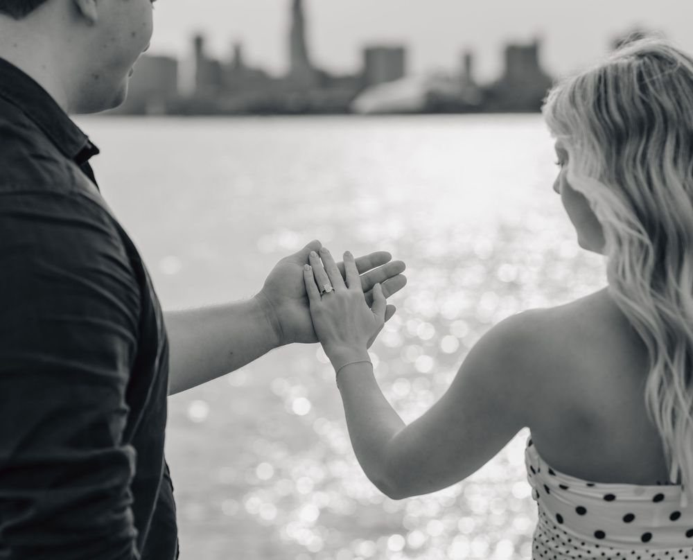 Black and white photo of a couple holding hands near a waterfront, with a city skyline in the background.