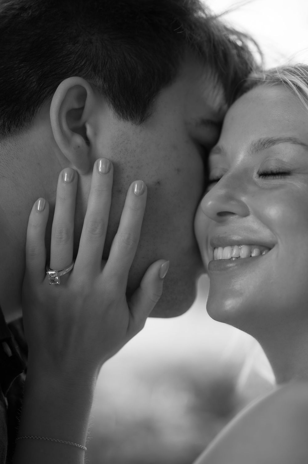 Black and white photo of smiling woman with engagement ring touching man's face as he kisses her cheek.