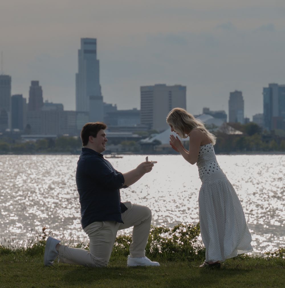Man proposes to woman on one knee by waterfront with city skyline backdrop on sunny day.