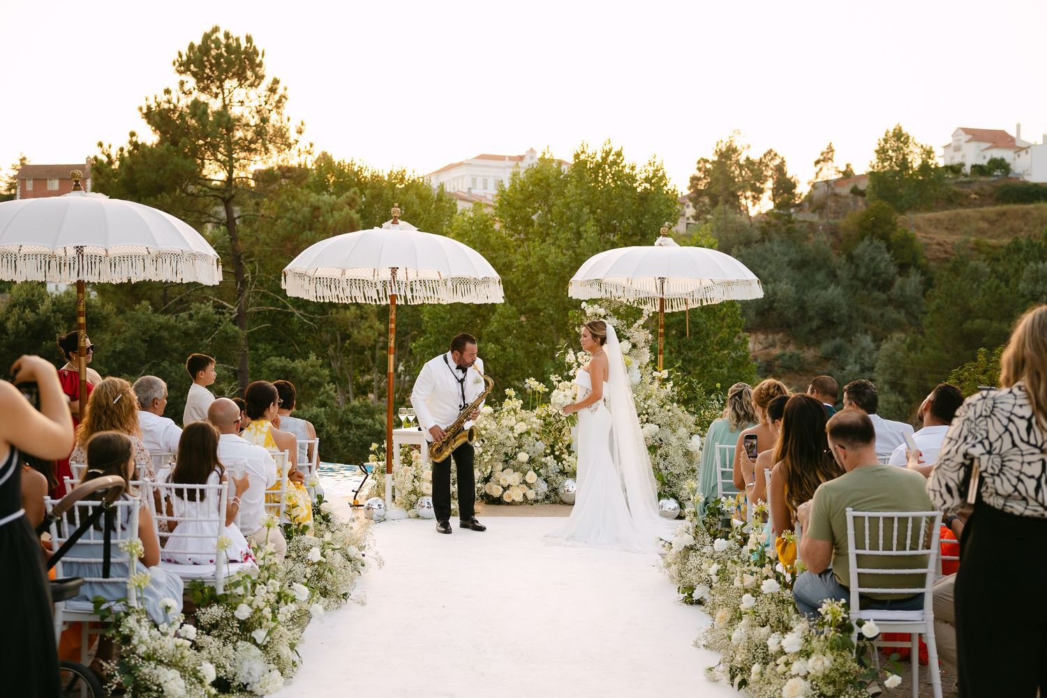 Groom plays saxophone to Bride at elegant outdoor wedding ceremony with white floral aisle, decorative umbrellas and lush greenery backdrop.