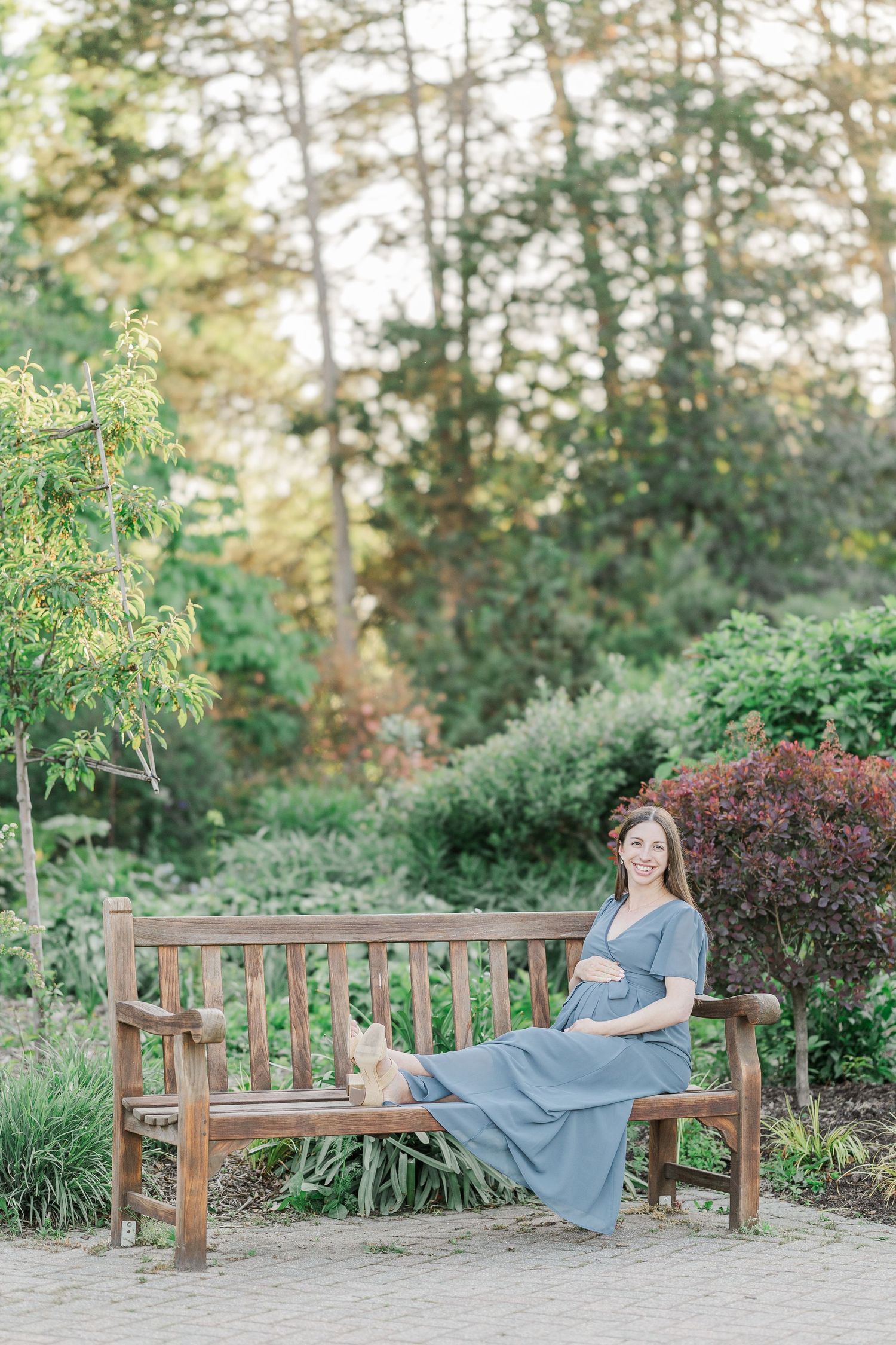 Pregnant woman in blue dress sitting on wooden bench in lush garden, smiling and cradling her baby bump.