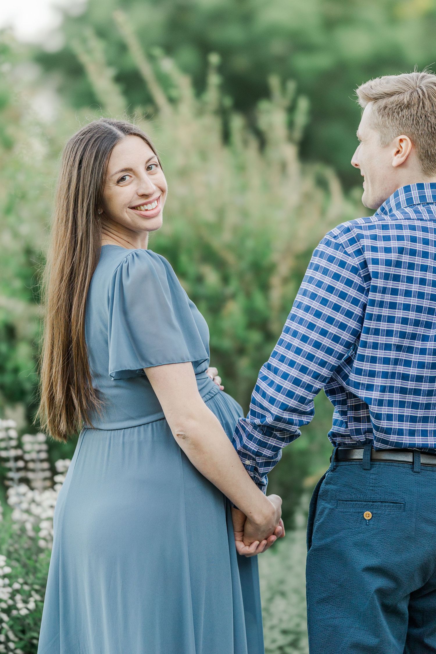 A smiling woman in a blue dress holds hands with a man in a plaid shirt outdoors.