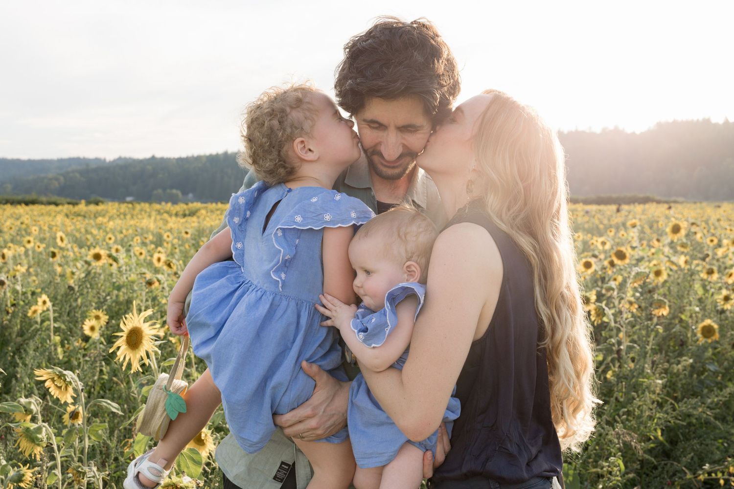 Family of four posing in a sunflower field at golden hour, parents kissing babies in matching blue dresses.