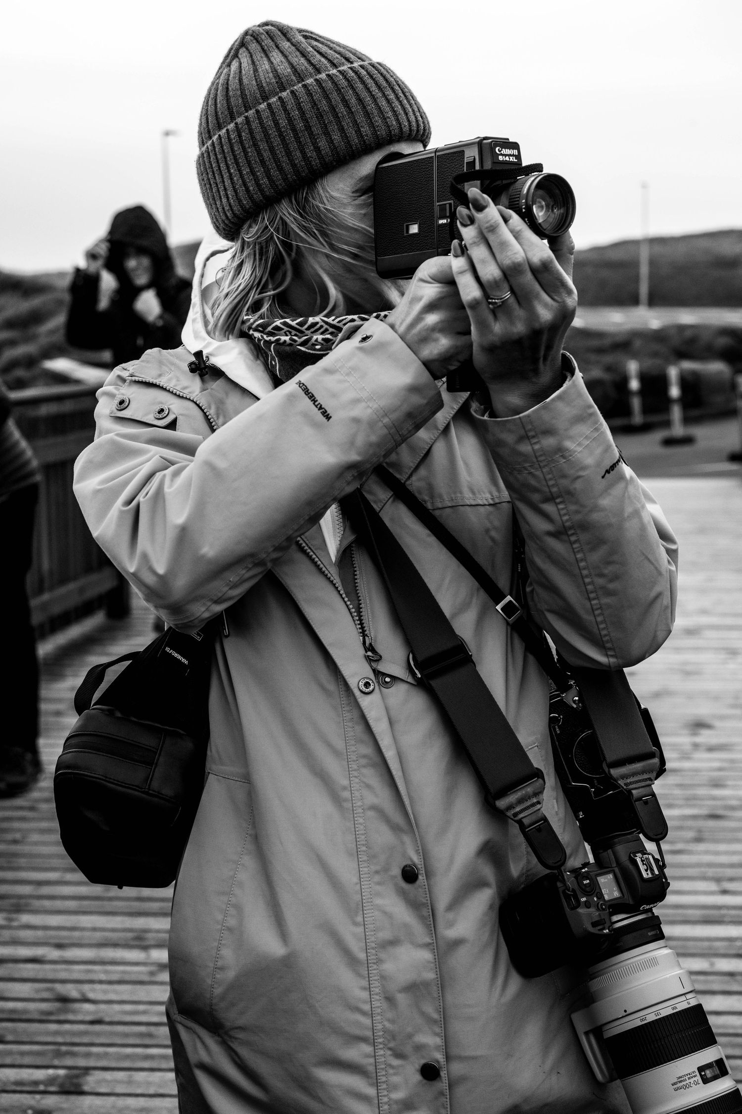 Black and white photo of a woman in a beanie and jacket photographing with a Canon camera, with a telephoto lens hanging at her side.