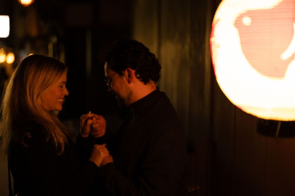 A couple holds hands intimately in a warmly lit setting with a glowing lantern in the background.