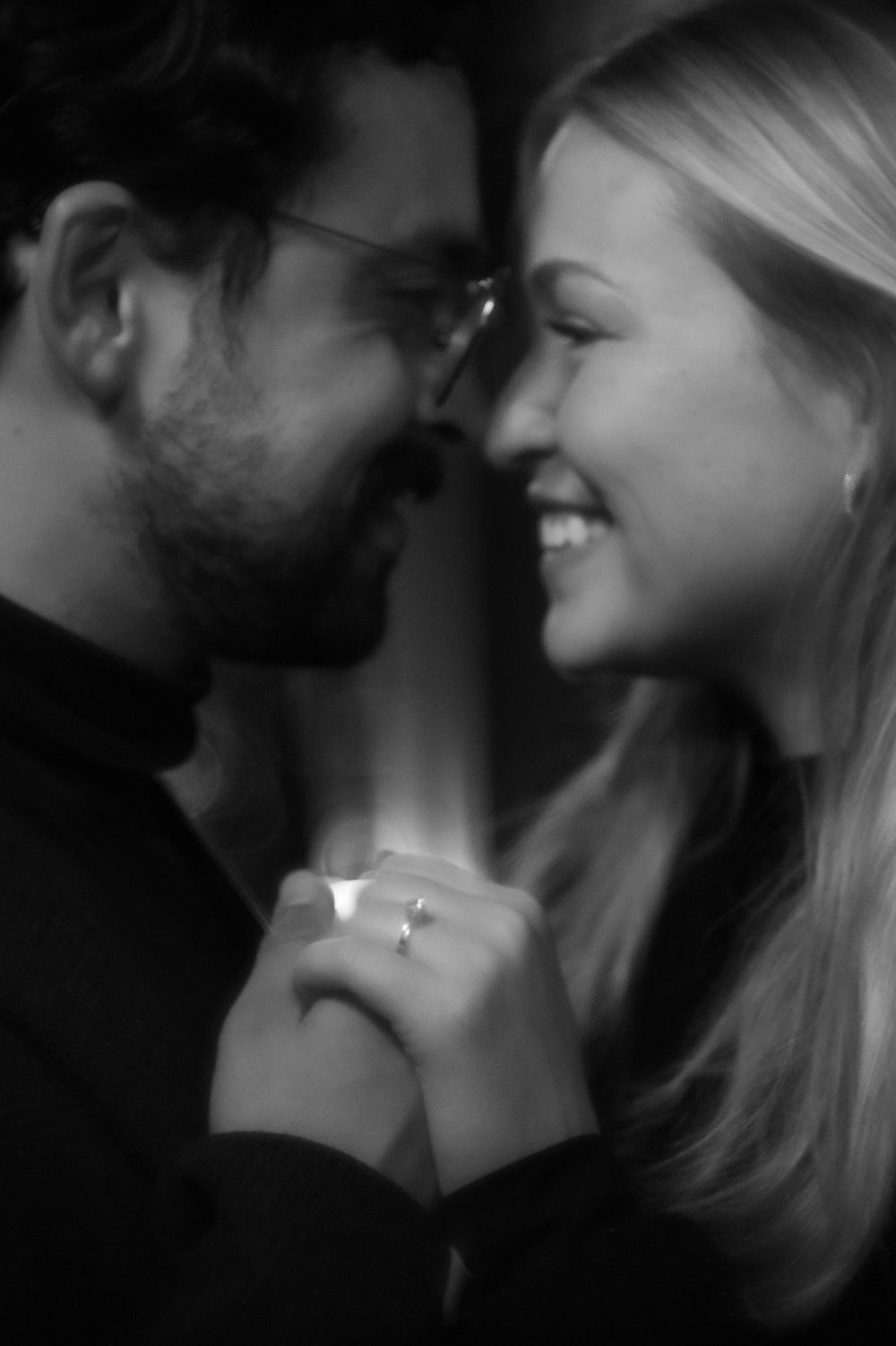 Black and white photo of a smiling couple holding hands closely, with an engagement ring visible.