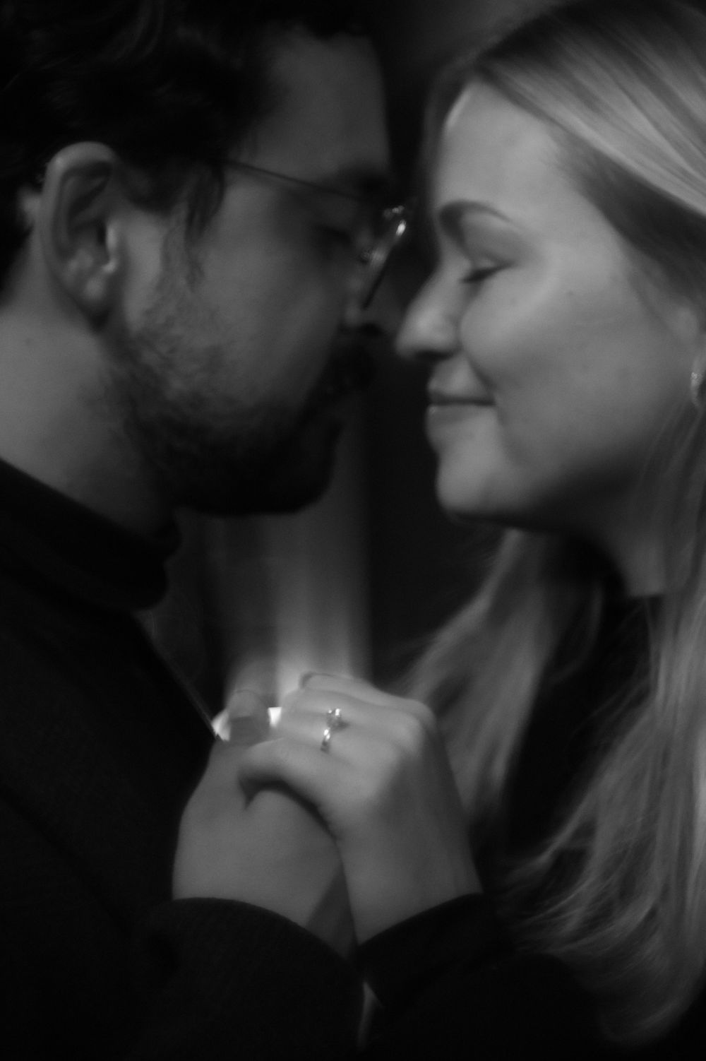 Black and white close-up of a couple holding hands and leaning in together, engagement ring visible.