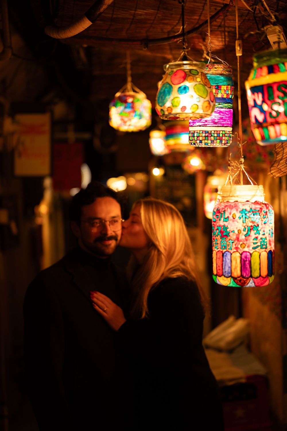 A woman kisses a man on the cheek beneath colorful decorative lanterns in a glowing night market.