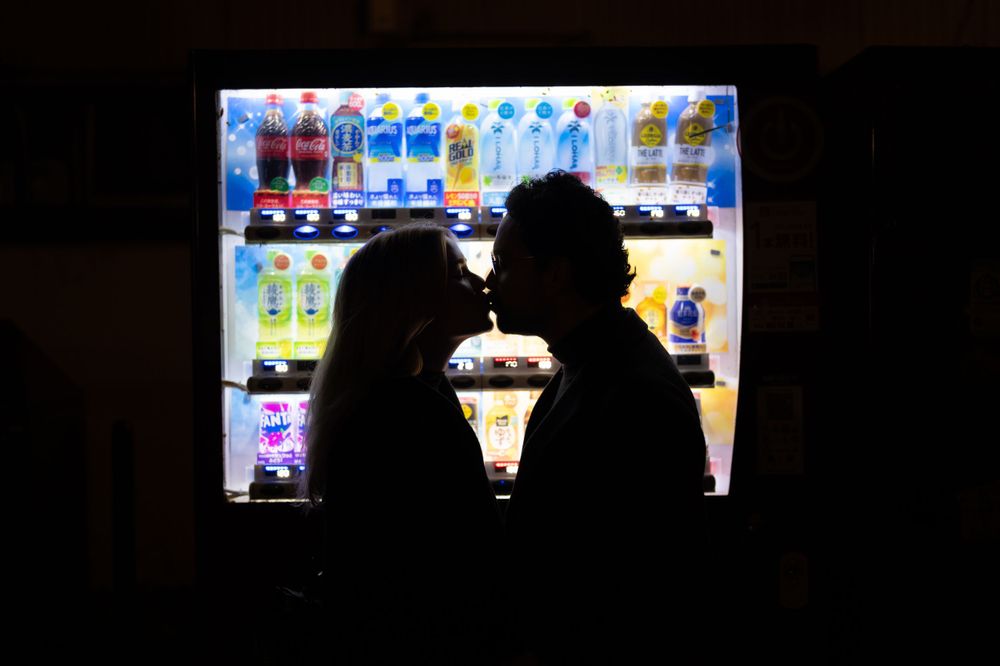 A couple shares a romantic kiss silhouetted against a brightly lit vending machine at night.
