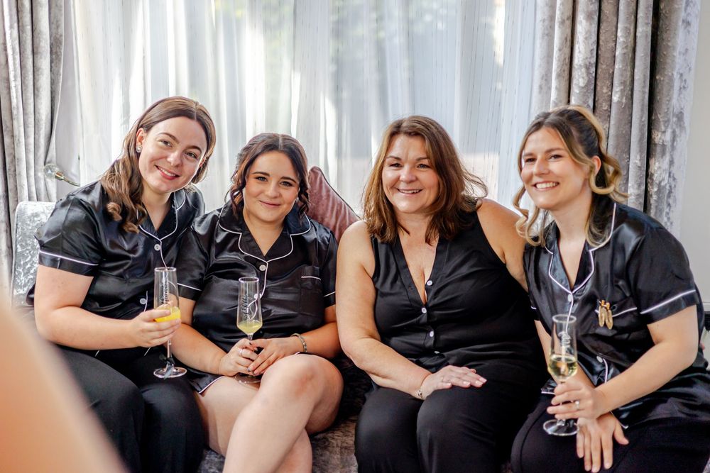 Four women in black pajamas smile and hold champagne glasses while sitting together on a couch at a bridal party.