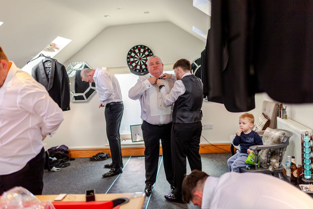 Groomsmen helping each other with ties in a bright attic room before a wedding.
