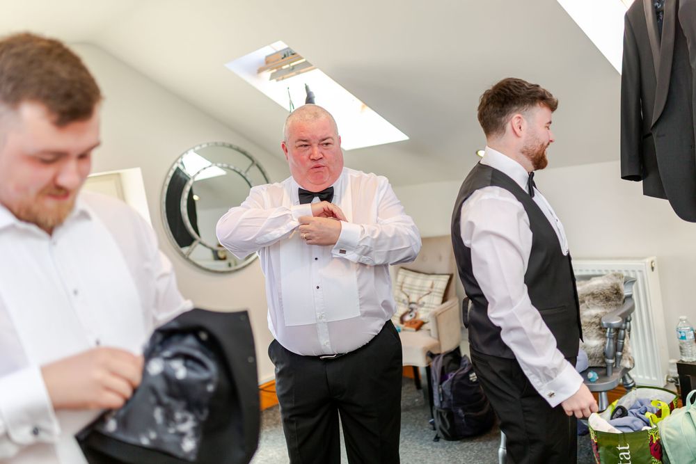 Three men in formal wedding attire adjusting bow ties and suits in a bright dressing room before the ceremony.