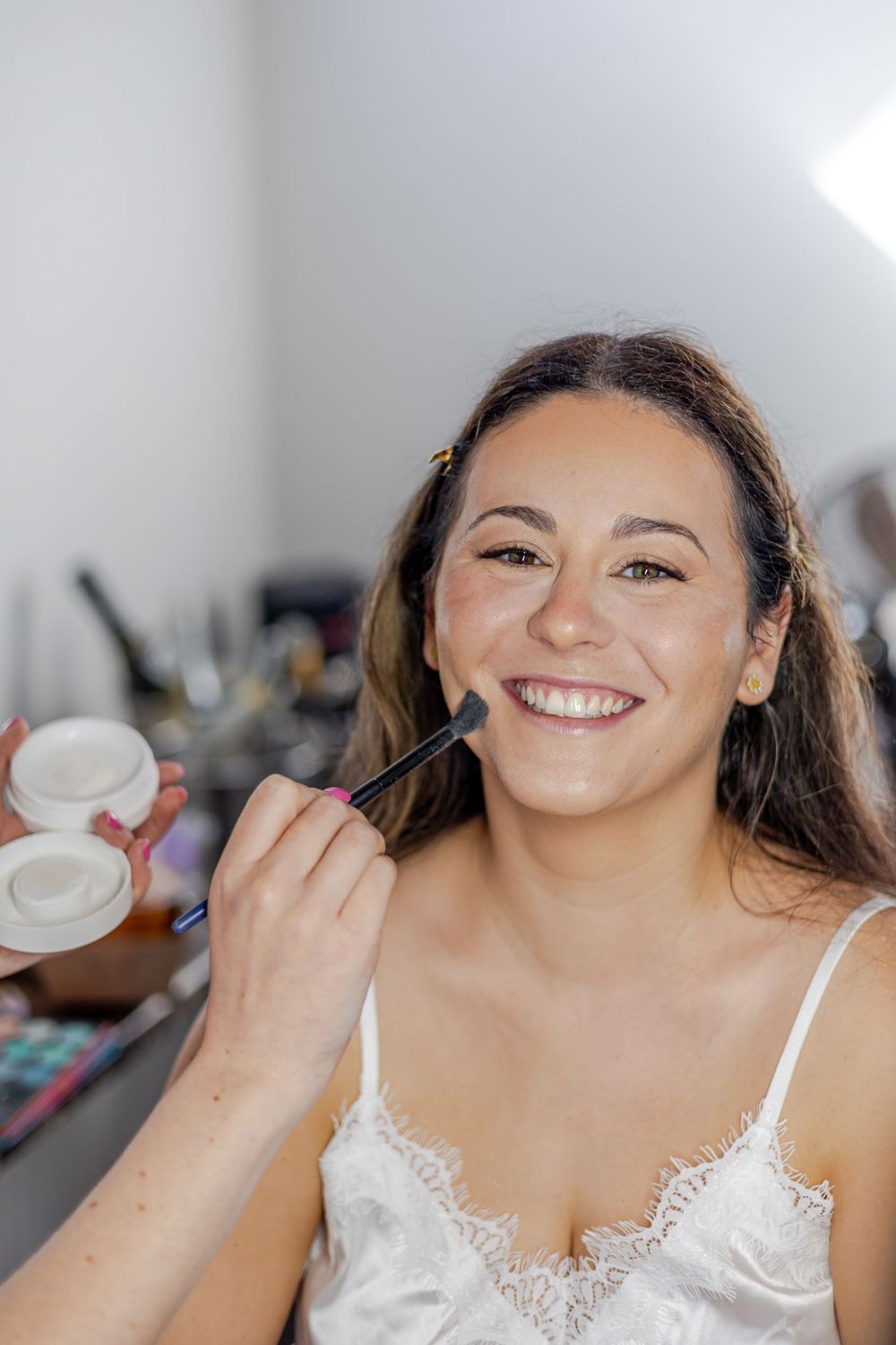 Smiling woman getting makeup applied with a brush while sitting in a beauty salon chair wearing a white lace top.