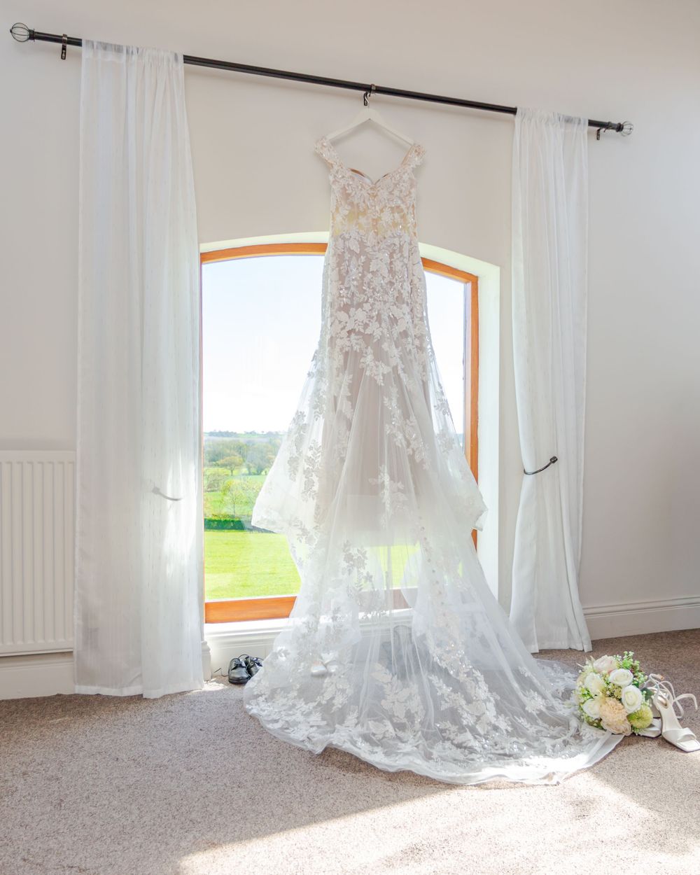 Elegant lace wedding dress with floral appliques and long train hanging by a sunlit window with white curtains.