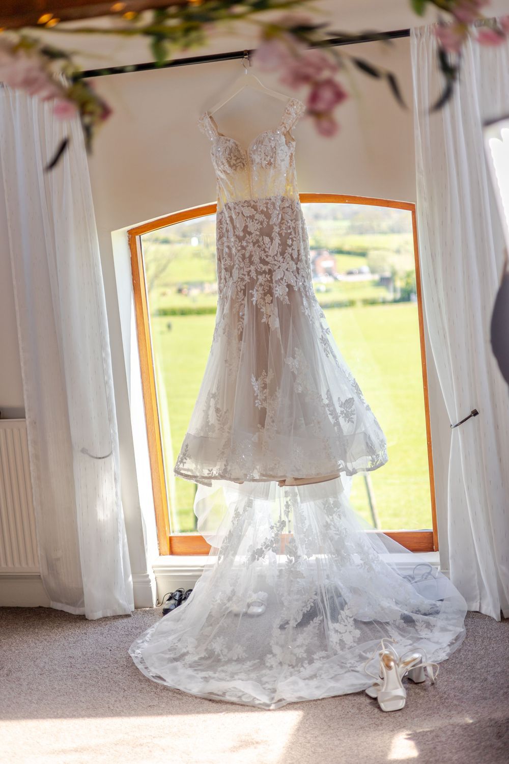 Elegant lace wedding dress hanging by a sunlit window with floral details and a flowing train, with bridal heels below.