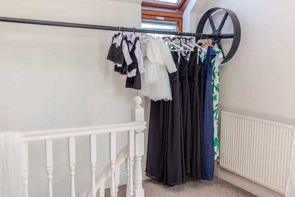 Formal dresses and suits hanging on a clothing rail near a staircase banister in a bright attic space.