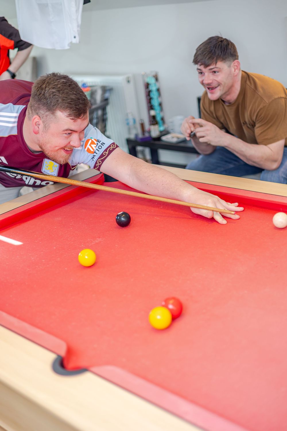 Two young men playing pool on a red billiard table, one leaning in to take a shot while the other watches.