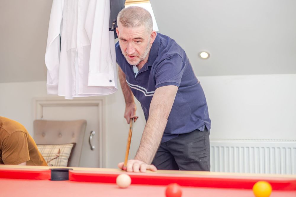 Elderly man leaning over a red pool table, carefully aiming his cue stick at billiard balls during a game.